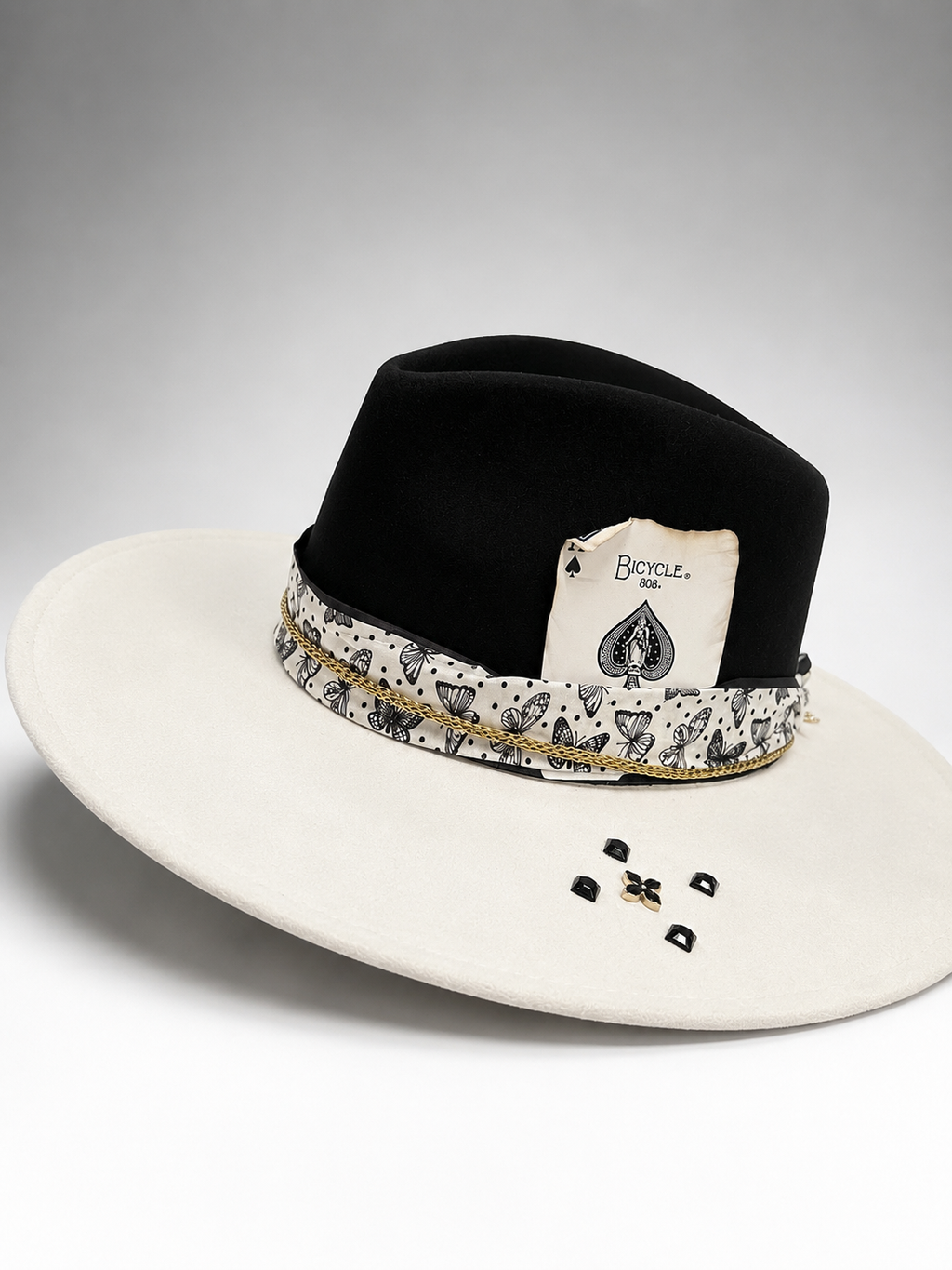 A black felt cowboy-style hat with a wide off-white brim, a patterned band featuring playing card suits, and a hanging tag that reads 'Bounty 1000'.