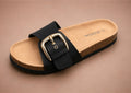 Birkenstock-style sandal with black strap and gold buckle on a beige background