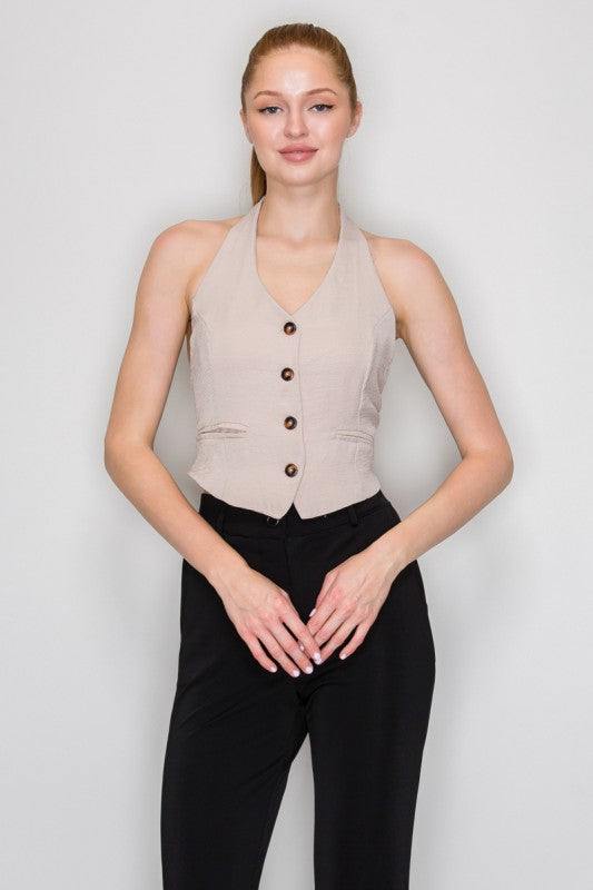 A woman modeling a sleeveless beige vest with dark buttons and black pants, standing against a light gray studio background.
