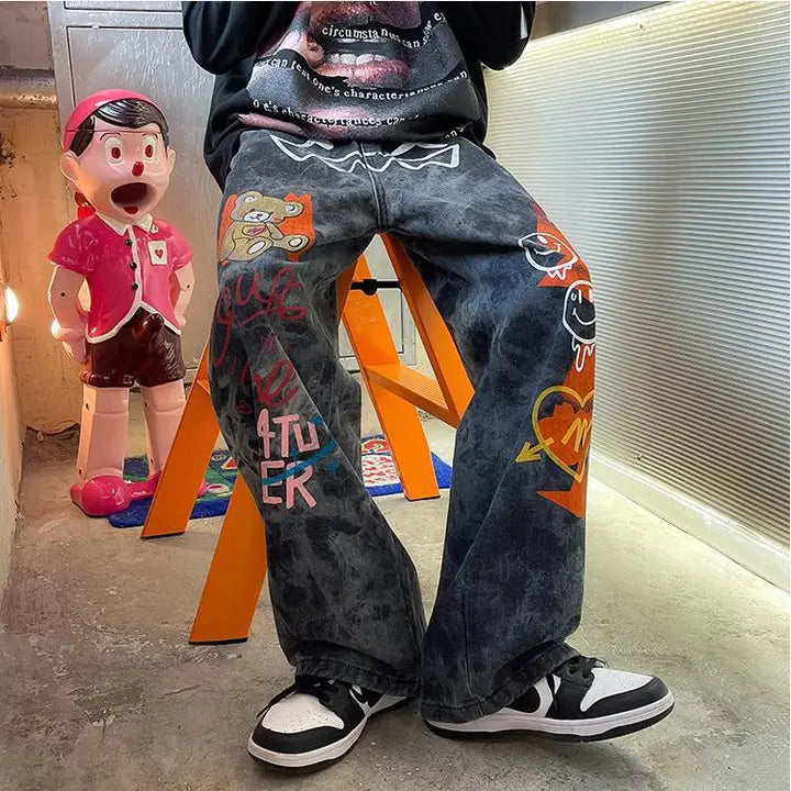 Person wearing black acid wash jeans with colorful graffiti designs including smiley face, teddy bear, and street art graphics on front legs on an orange chair. 