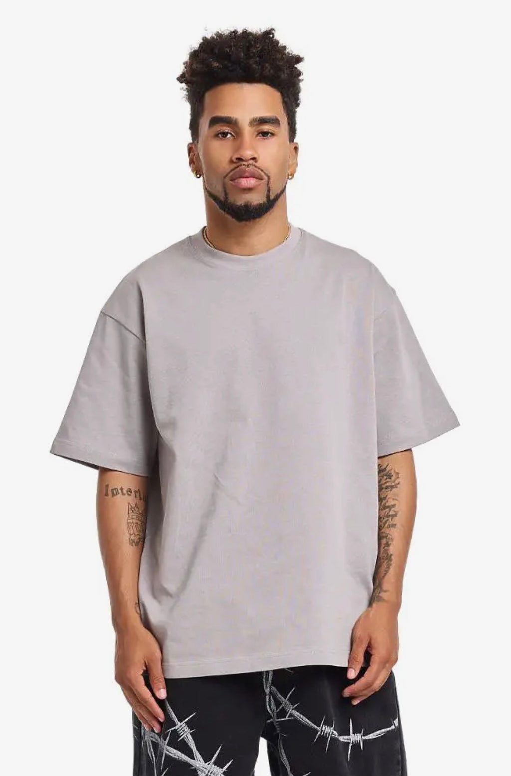 A man wearing an oversized light gray crewneck t-shirt and black barbed wire print pants, standing against a plain light gray background.