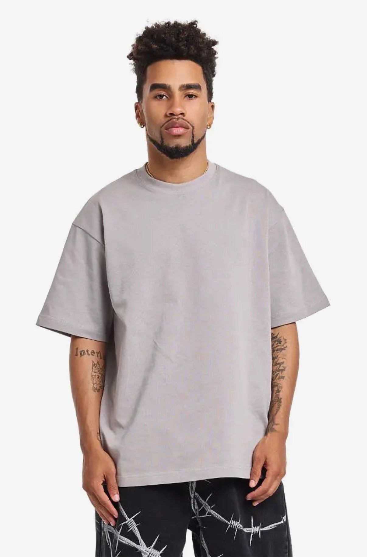 A man wearing an oversized light gray crewneck t-shirt and black barbed wire print pants, standing against a plain light gray background.