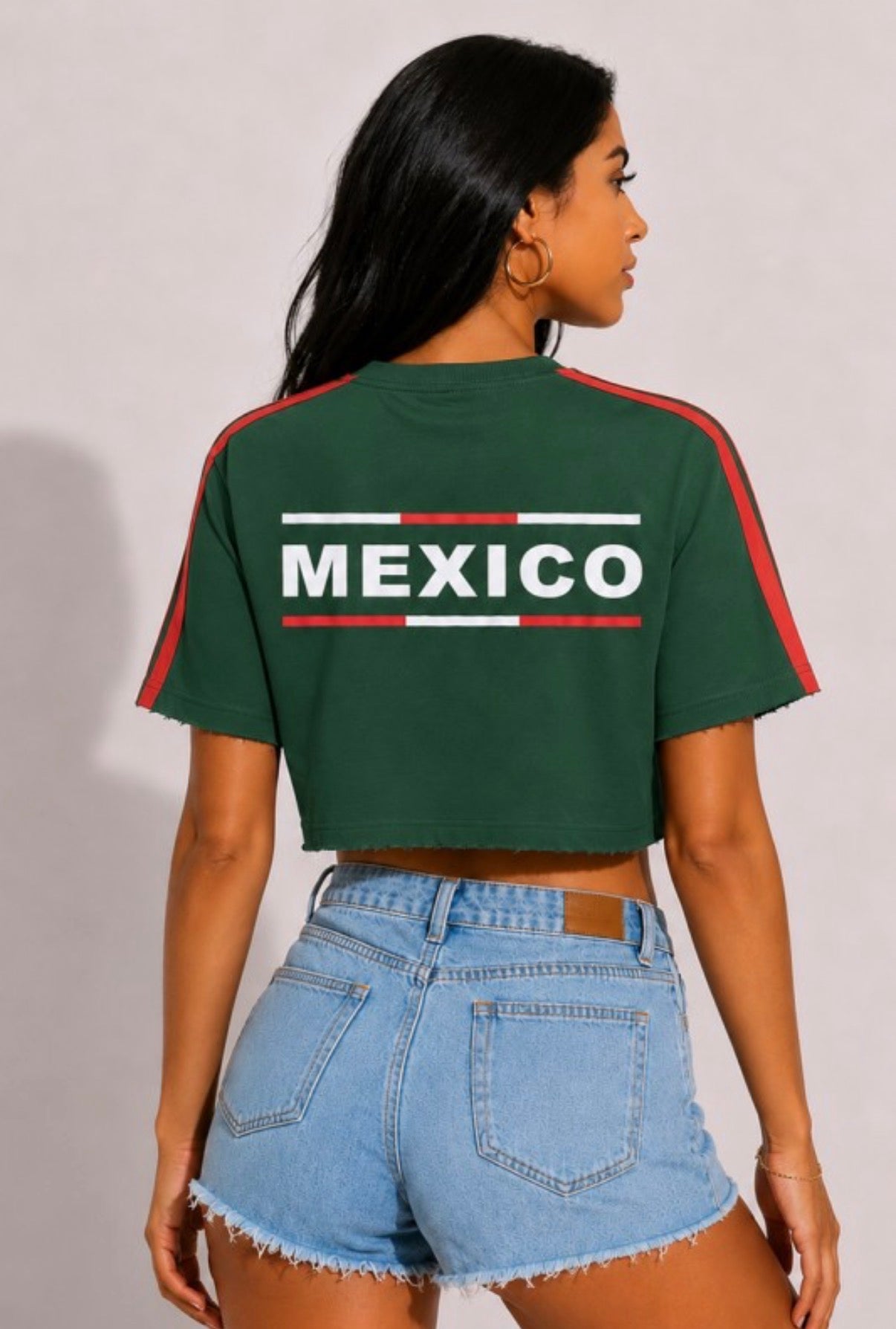 A woman shown from the back wearing a dark green cropped t-shirt with 'MEXICO' printed in white across the back, flanked by red and white stripes, paired with light blue denim shorts.