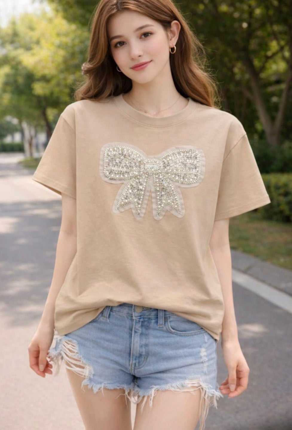 A woman wearing a beige short-sleeve t-shirt with a large sparkling bow applique on the front, paired with light blue denim shorts, standing outdoors on a paved path with trees in the background.