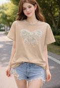 A woman wearing a beige short-sleeve t-shirt with a large sparkling bow applique on the front, paired with light blue denim shorts, standing outdoors on a paved path with trees in the background.