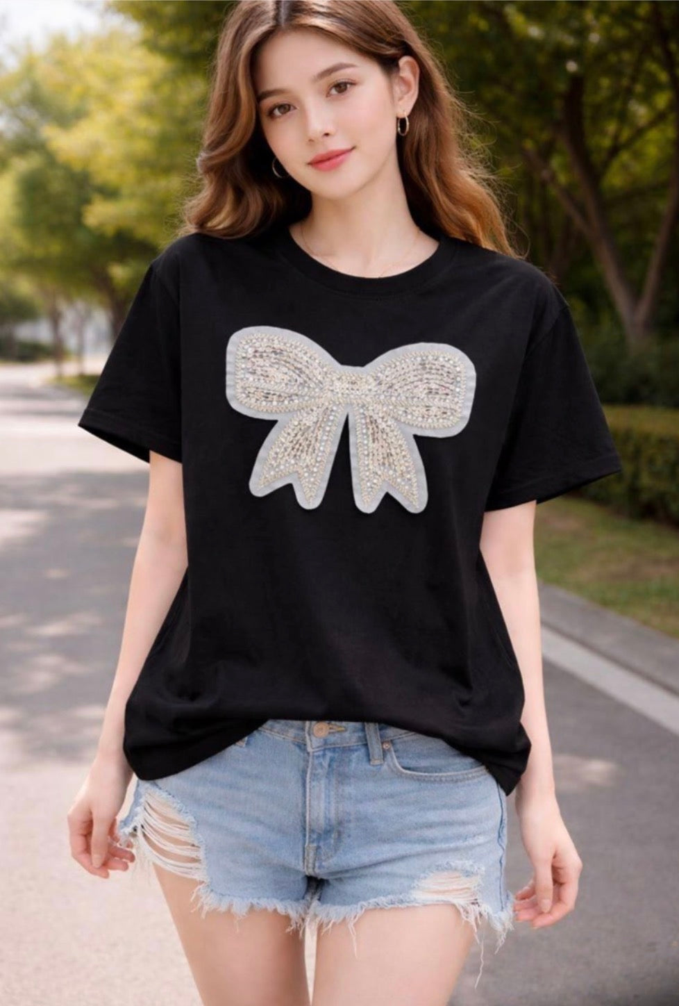 A woman wearing a black short-sleeve t-shirt with a large, sparkling silver bow graphic on the front, paired with light-wash distressed denim shorts, standing outdoors on a paved path with trees in the background.