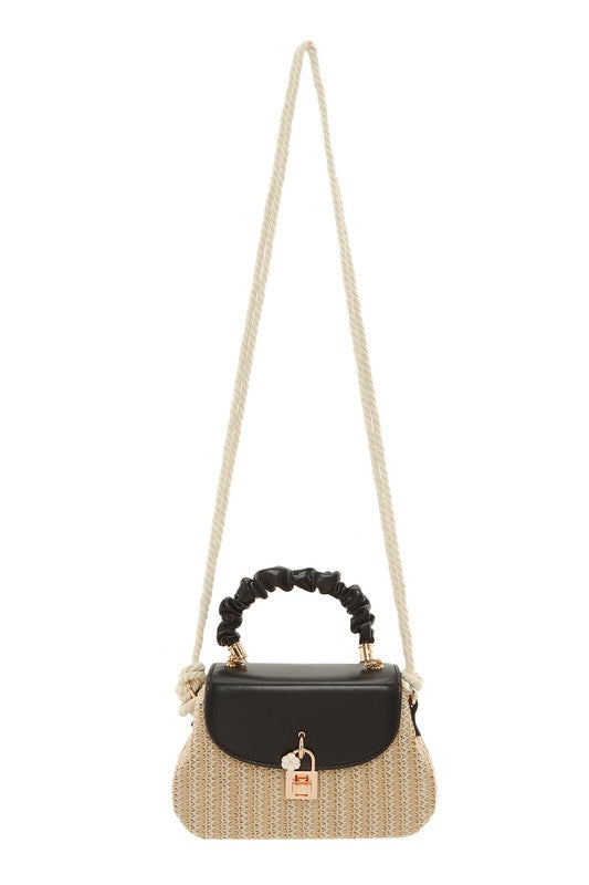Small woven shoulder bag featuring a natural straw-textured body with a black flap closure and gold-tone clasp detail. The bag has a ruched black top handle with gold hardware accents and a long cream-colored rope shoulder strap. Displayed against a light gray background, the design combines a coastal, woven look with elegant black-and-gold details.