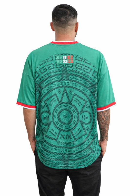 Person wearing a green t-shirt with a geometric pattern on the back, standing against a white background.