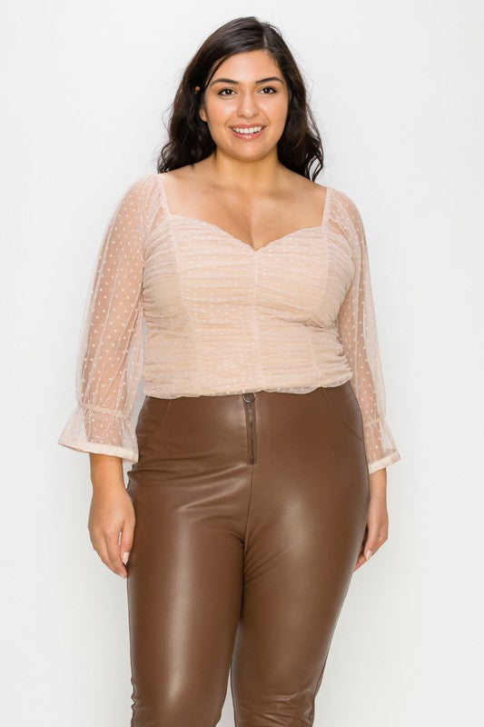 A woman modeling a beige sheer long-sleeve crop top with a sweetheart neckline, paired with brown faux-leather pants, against a plain white studio background.