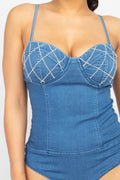 A close-up of a model wearing a blue denim one-piece swimsuit with white rhinestone crisscross detailing on the bust cups, set against a plain light gray background.