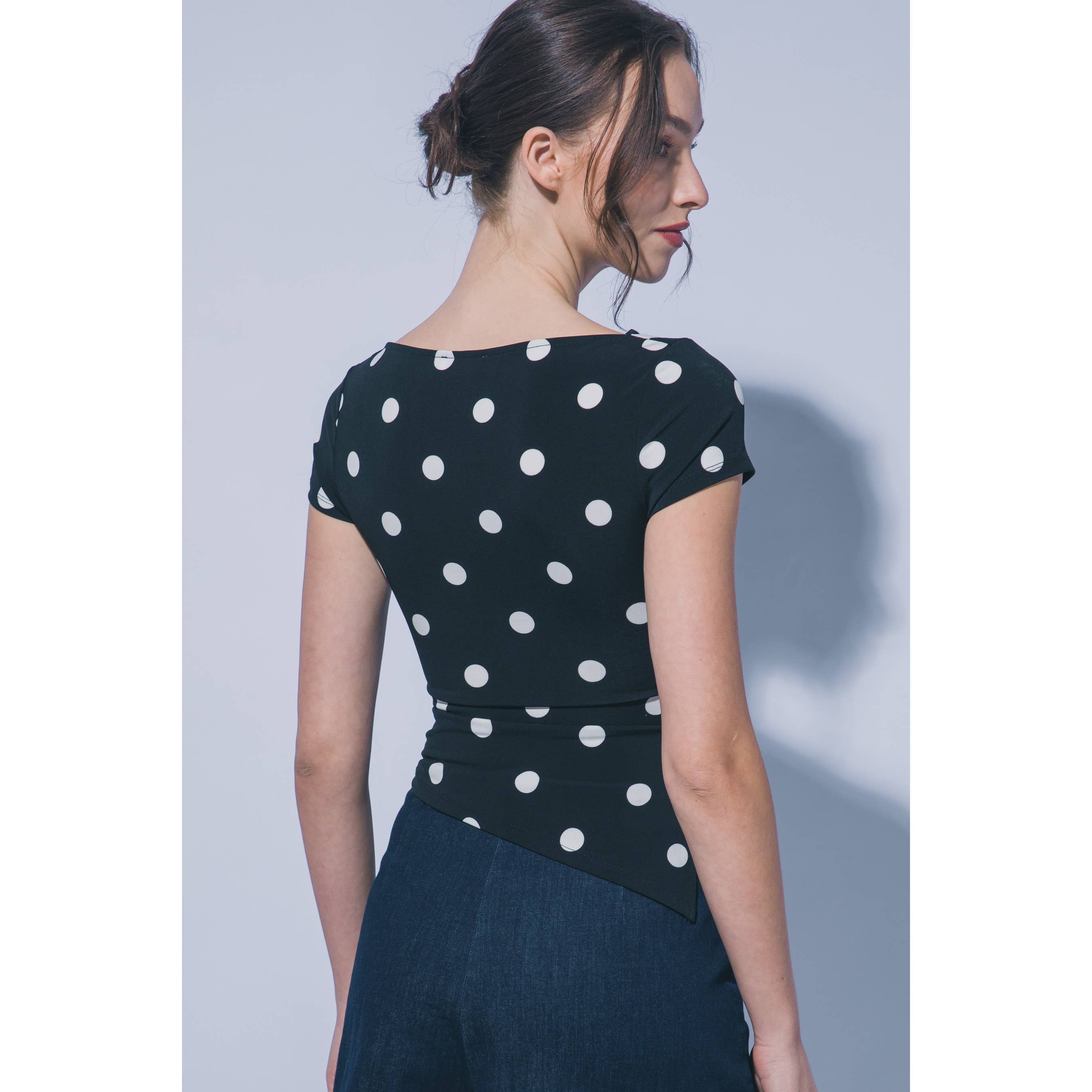 A woman shown from the back wearing a black short-sleeve top with white polka dots and a dark blue skirt, set against a plain light gray studio background.