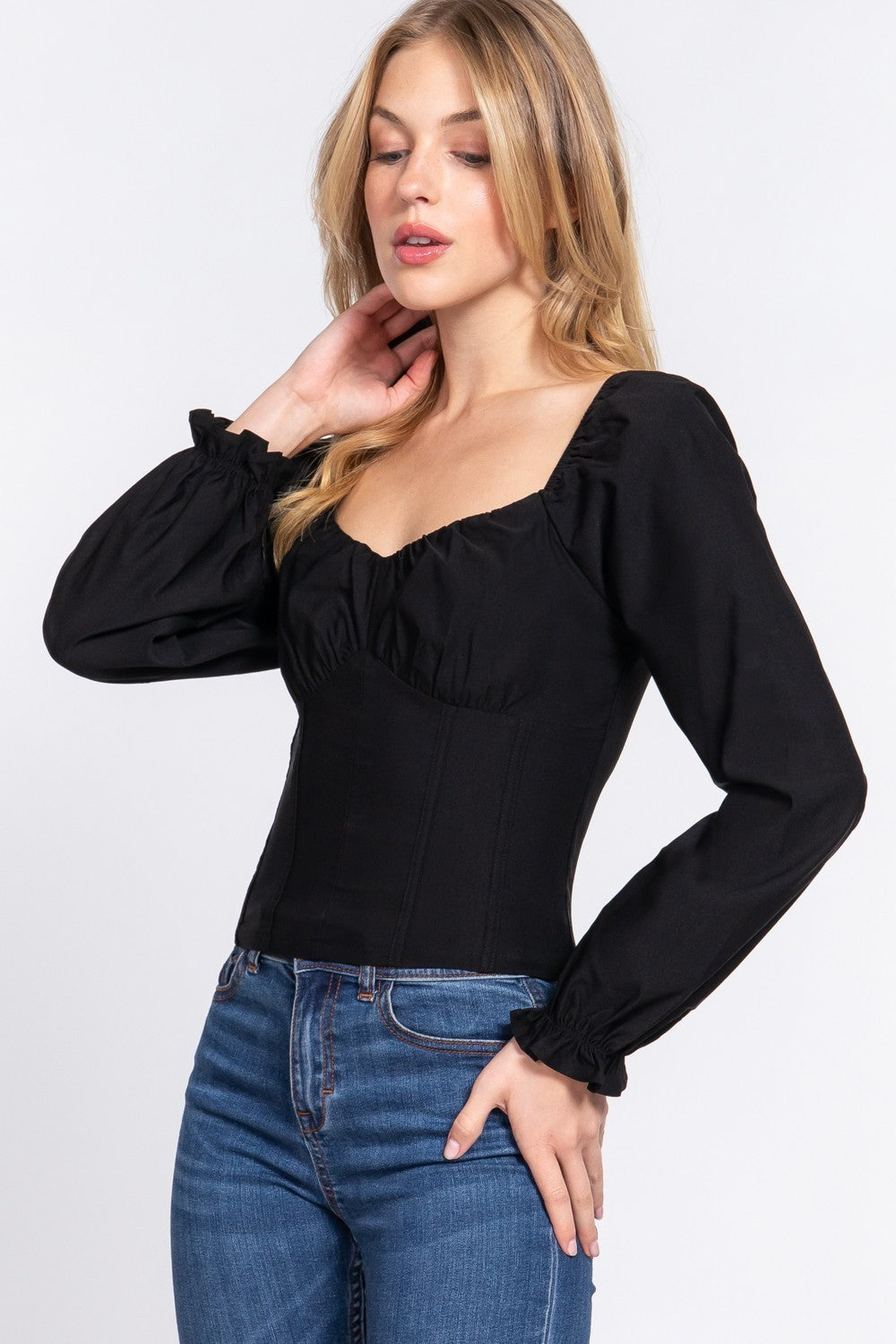 A woman wearing a black long-sleeve blouse with a square neckline, gathered puff sleeves, and ruffled cuffs, paired with blue jeans, against a light gray studio background.