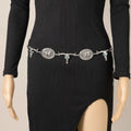 A silver western-style belt with three large engraved oval medallions and hanging skull charms, shown worn at the waist on a black ribbed long-sleeve dress displayed on a mannequin.