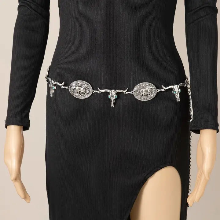 A silver western-style belt with three large engraved oval medallions and hanging skull charms, shown worn at the waist on a black ribbed long-sleeve dress displayed on a mannequin.