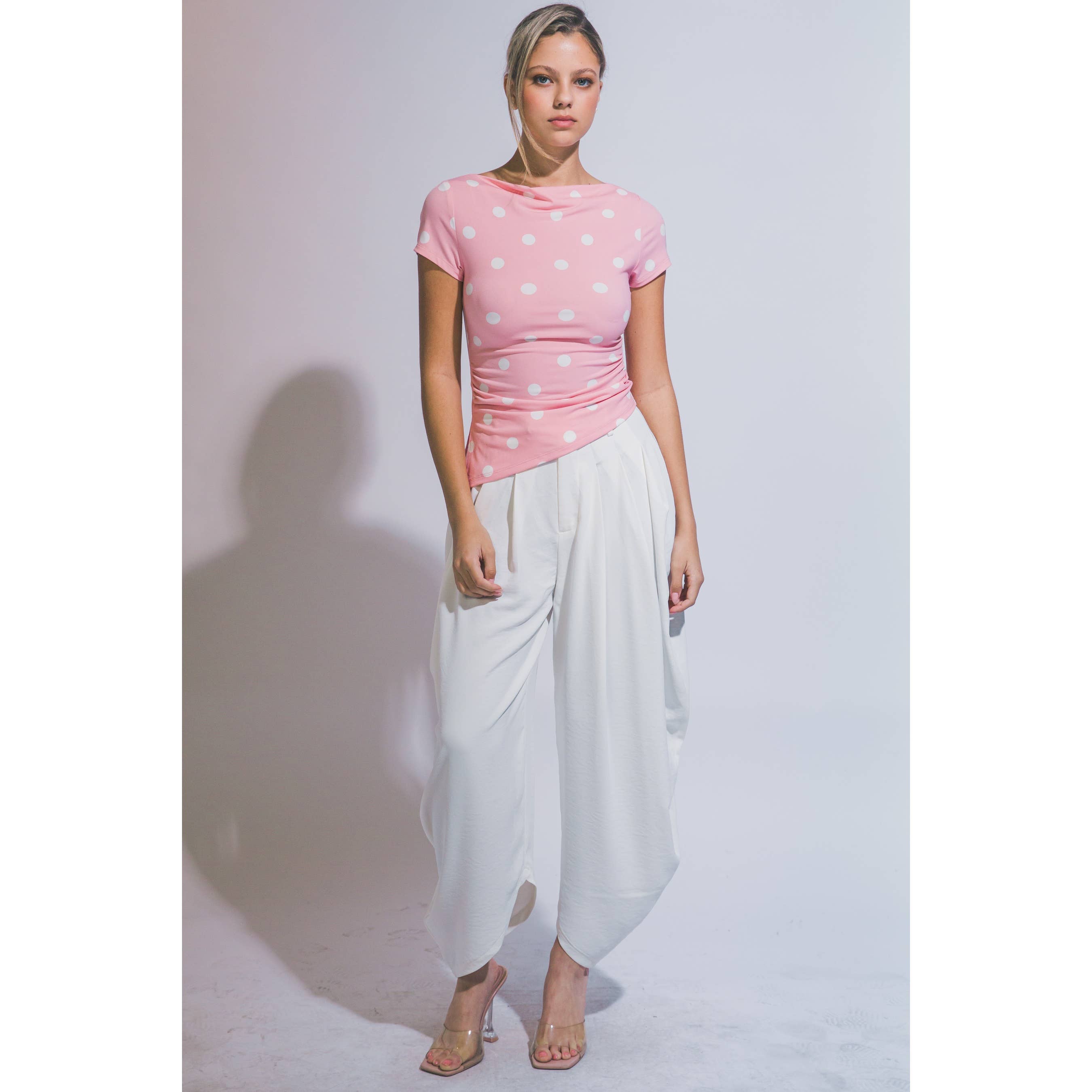 A woman modeling a pink short-sleeve top with white polka dots paired with loose white draped pants, standing against a light gray studio background.