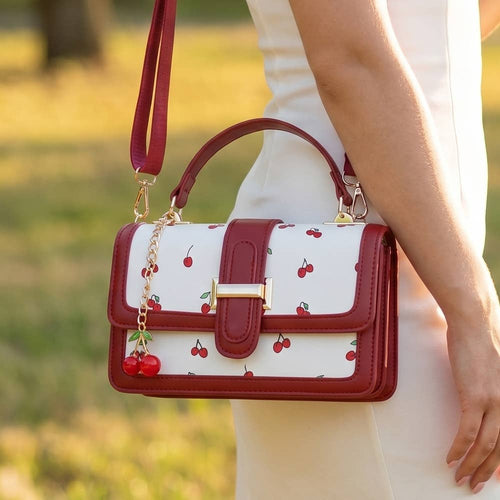 A woman in a white dress carries a cherry-patterned handbag with a white body, deep red trim, a top handle, and a shoulder strap, photographed outdoors with a blurred grassy background.