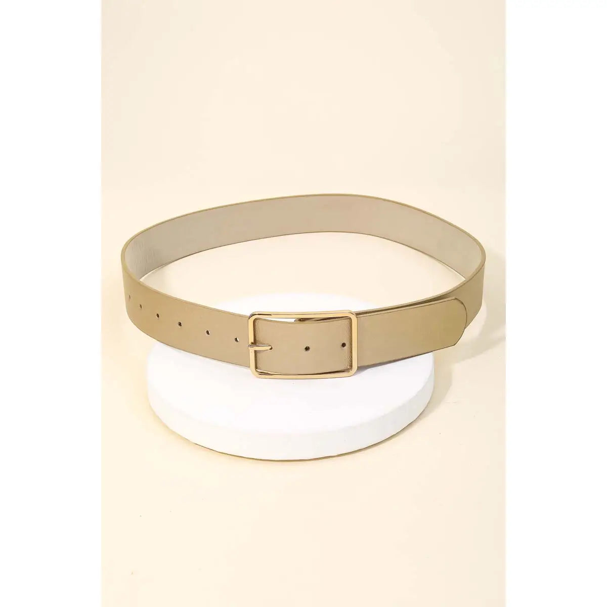 A slim beige belt with a gold-tone rectangular buckle, displayed on a white circular pedestal against a light beige background.
