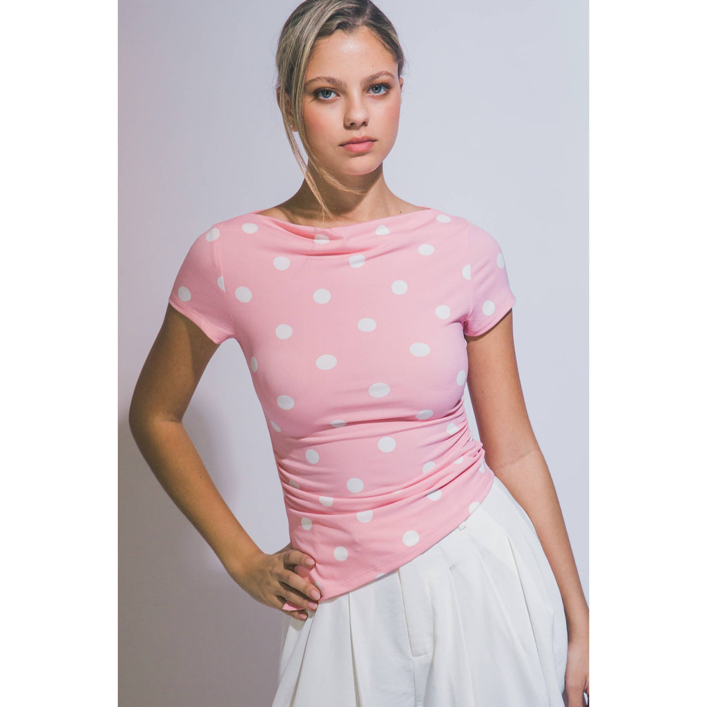 A woman wearing a pink short-sleeve top with white polka dots and a gathered waist, paired with a white skirt, posing against a light gray studio background.