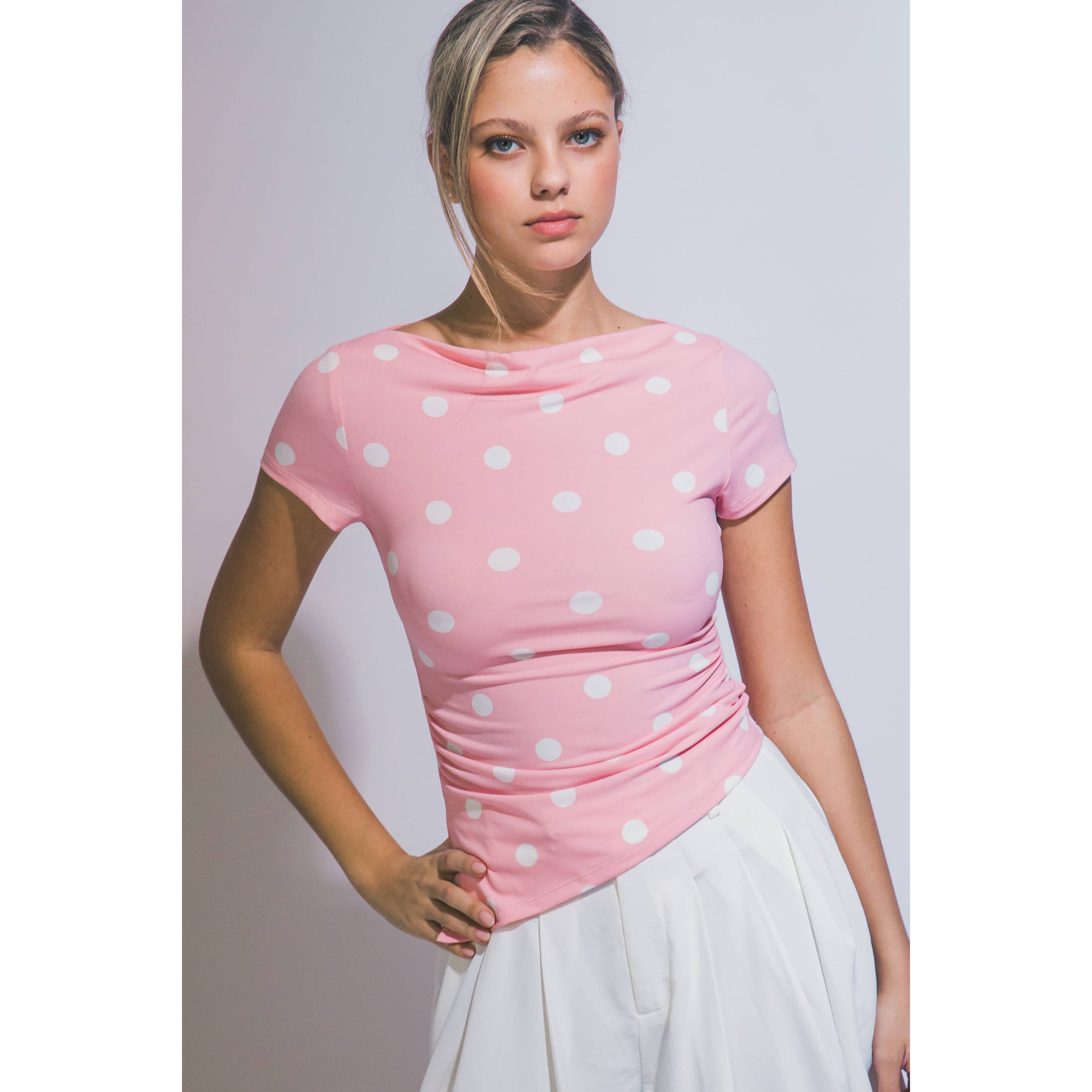 A woman wearing a pink short-sleeve top with white polka dots and a gathered waist, paired with a white skirt, posing against a light gray studio background.