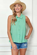 A woman wearing a mint green sleeveless button-up shirt and denim shorts, standing against a white paneled wall and wearing a straw hat.