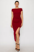A woman modeling a deep red off-the-shoulder midi dress with a ruched bodice and a high side slit, paired with black strappy high-heeled sandals against a plain white studio background.